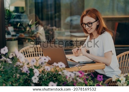 Redhead skillful woman student writes in notebook sits in front of opened laptop computer notes information for planning project does remote job poses in outdoor cafe uses modern technologies