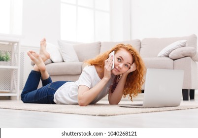 Redhead smiling student girl using laptop for studying and chatting on cell phone while lying on the floor carpet. Technology, communication and coziness concept - Shutterstock ID 1040268121