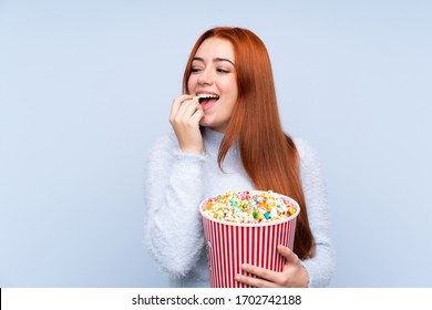 Redhead teenager girl over isolated blue background holding a big bucket of popcorns