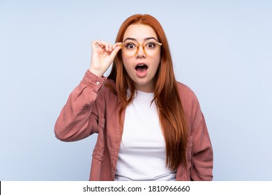 Redhead teenager girl over isolated blue background with glasses and surprised