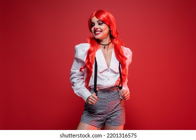 Redhead Woman With Clown Makeup Posing In White Blouse And Shorts With Suspenders Isolated On Red