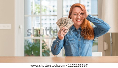 Redhead woman holding dollar bank notes at home with happy face smiling doing ok sign with hand on eye looking through fingers