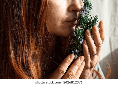 Redhead Woman holding in her hands a bunch of blooming rosemary and inhales it's scent. Herbalist woman preparing fresh organic herbs for natural herbal methods of treatment. Alternative medicine.