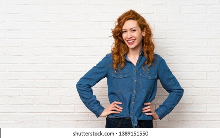 Redhead woman over white brick wall posing with arms at hip and smiling