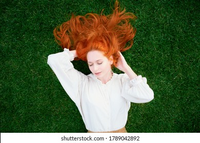 Redhead Woman Is Resting On Bright Green Lawn, Her Eyes Closed And Dreaming. Model With Long Thick Hair Enjoys Summer