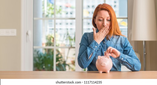 Redhead woman saves money in piggy bank at home cover mouth with hand shocked with shame for mistake, expression of fear, scared in silence, secret concept Stock Photo