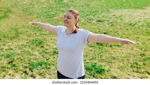 Redhead woman standing with arms outstretched closed eyes smiles feels love to life, welcoming new good day. Fitness female doing warmup exercise on nature background