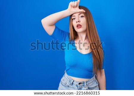 Redhead woman standing over blue background making fun of people with fingers on forehead doing loser gesture mocking and insulting. 