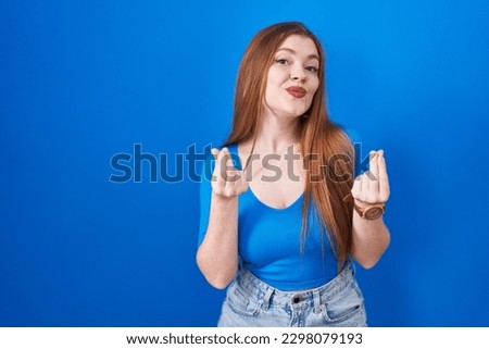 Redhead woman standing over blue background doing money gesture with hands, asking for salary payment, millionaire business 