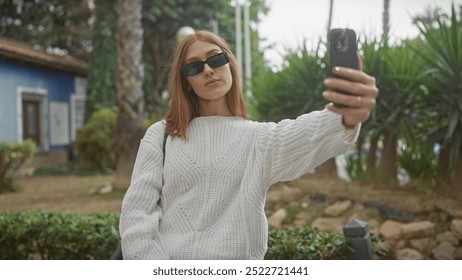 A redhead woman in sunglasses captures a selfie in a park wearing a white sweater for a casual outdoor portrait under greenery. - Powered by Shutterstock