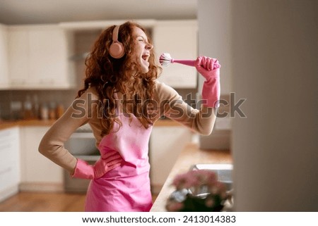 Redhead woman washing dishes in the kitchen and listening to music by wireless headphones, happy   female cleaning plate with detergent, enjoying making domestic chores