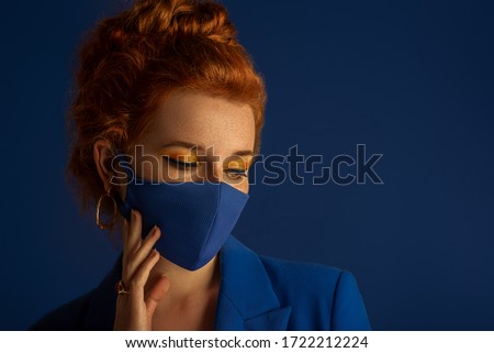 Redhead woman wearing trendy fashion blue monochrome outfit with luxury designer protective face mask. Model has matching bold eyes makeup. Vogue, style during quarantine of coronavirus outbreak. 