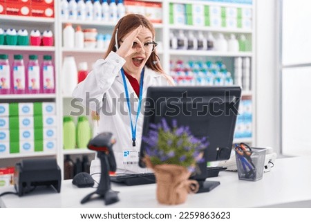 Redhead woman working at pharmacy drugstore wearing headset smiling happy doing ok sign with hand on eye looking through fingers 