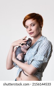 Redhead Young Woman Gently Hugging Canadian Sphynx Cat, Looking At Camera. Studio Shot On White Background. Portrait Caucasian Hipster With Short Hair Dressed In Striped White-blue Shirt. Part Series.