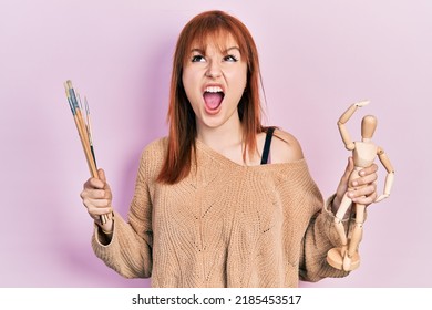 Redhead young woman holding small wooden manikin and painter brushes angry and mad screaming frustrated and furious, shouting with anger looking up.  - Powered by Shutterstock