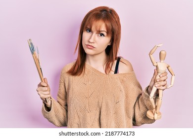 Redhead young woman holding small wooden manikin and painter brushes skeptic and nervous, frowning upset because of problem. negative person. 