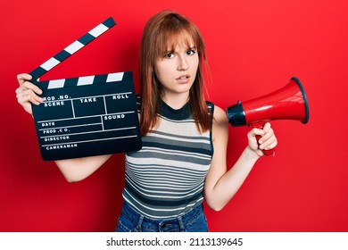 Redhead Young Woman Holding Video Film Clapboard And Megaphone Clueless And Confused Expression. Doubt Concept. 