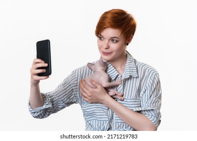 Redhead Young Woman Hugging Sleeping Domestic Kitten, Using Her Cell Phone And Taking Selfie At Smartphone Camera. Studio Shot On White Background. Part Of Series. Facial Expression, Positive Emotion