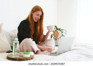 Redhead young woman sitting in bedroom and doing online shopping. Girl looking at laptop and buying natural beauty cosmetic products by the internet at home. Foto Stock