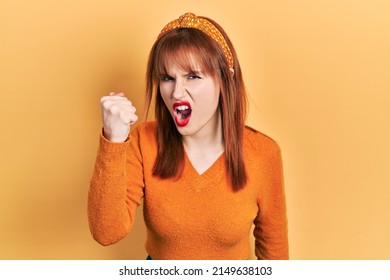 Redhead young woman wearing casual orange sweater angry and mad raising fist frustrated and furious while shouting with anger. rage and aggressive concept. 