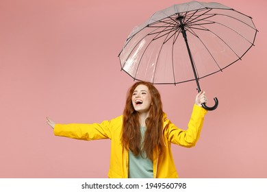 Redhead young woman in yellow waterproof raincoat hood outerwear hold transparent umbrella outstretched hand isolated on pastel pink background studio Outdoor lifestyle wet fall weather season concept