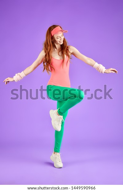 red-headed girl with freckles in pink bodysuit,\
green leggins and pink sun cap doing aerobics exercises keeping one\
leg raised and looks down with smile, enjoys training. 80s style\
aerobics concept