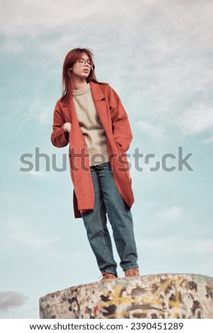 Redheaded teenage girl in red coat with hands in pockets standing on concrete podium against sky. Low angle view.