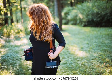 Redheaded Woman Wearing All Black Clothes And A Leather Camera Harness With Camera And Bag While Standing In Green Grass And Sunlight