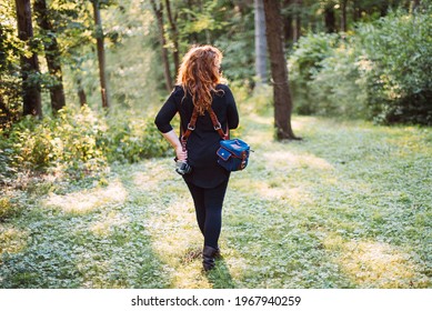 Redheaded Woman Wearing All Black Clothes And A Leather Camera Harness With Camera And Bag While Standing In Green Grass And Sunlight