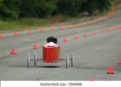 Red/Orange Soap Box Race Car Speeds Down The Track. Shallow Depth Of Focus On Car And Driver While Forest Background Becomes A Blur.