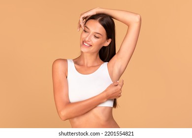 Reducing body odor. Happy fit lady standing with one hand up, demonstrating her smooth depilated armpit, posing in white top over beige background