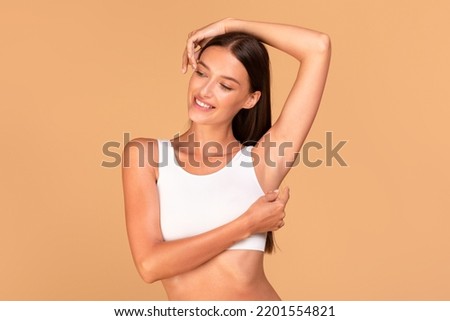 Reducing body odor. Happy fit lady standing with one hand up, demonstrating her smooth depilated armpit, posing in white top over beige background