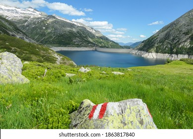 A red-white-red path mark on a lush green meadow in Austrian Alps. There is an artificial lake, shining with navy blue color. The dam is surrounded by high mountains. Controlling the nature