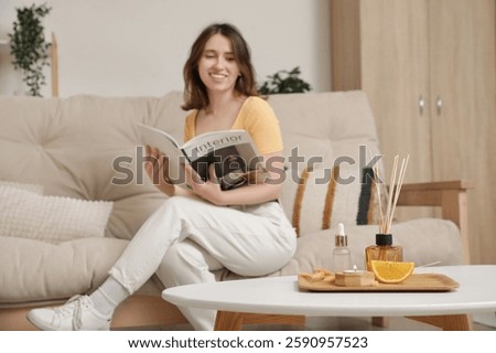 Reed diffuser with orange piece, essential oil and burning candle on table against woman reading magazine at home, closeup