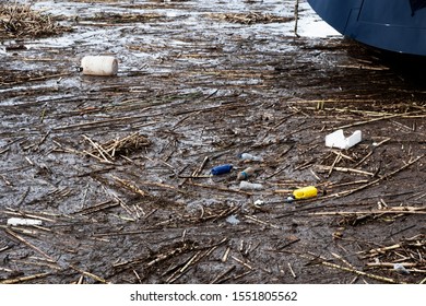 Reeds, Plastic Bottles And Other Garbage Floating On The Surface Of The Water In A Port Or In A Pond Or A Lake, Next To The Hull Of A Boat