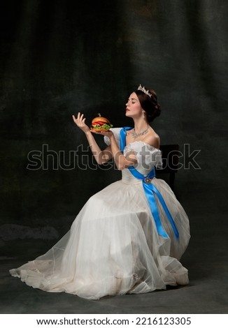 Refined taste. Young beautiful queen or princess in white medieval outfit eating burger on dark background. Concept of comparison of eras, fast food, fashion, beauty and ad