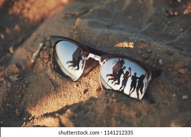 Reflecting Sunglasses On A Sandy Beach In The Summer, And The Blue Sky With People Is Reflected In Glasses