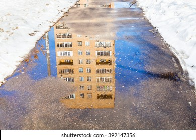 Reflection Of Apartment House In Puddle Of Melting Snow On Urban Footpath In Sunny Spring Day