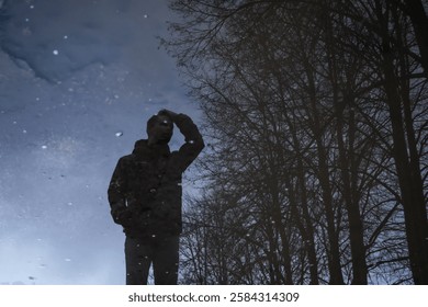 A man’s reflection appears in a puddle, blending with bare trees and sky. His posture suggests deep thought, creating a surreal and introspective mood with nature and urban elements intertwined