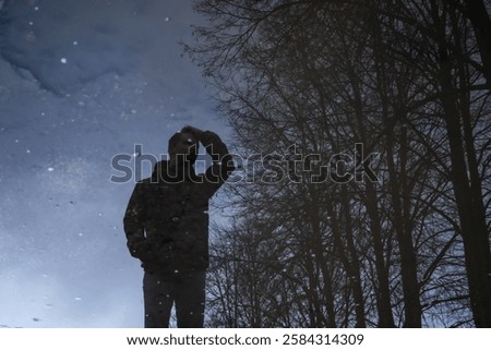 A man’s reflection appears in a puddle, blending with bare trees and sky. His posture suggests deep thought, creating a surreal and introspective mood with nature and urban elements intertwined