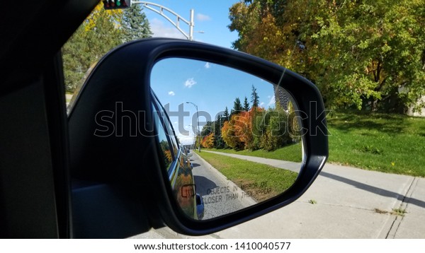 Reflection of asphalt highway road with cars\
and trees at the car side mirror. The road in car mirror with blue\
sky and green, orange trees in the\
background.