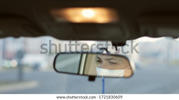 Reflection in back view mirror of woman in\
protective medical mask. Driving car through city during outbreak\
of dangerous disease around world. Covid\
-19.