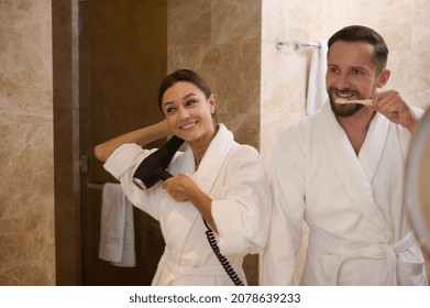 Reflection In Bathroom Mirror Of Happy Young Heterosexual Couple In Love During Morning Hygiene. Handsome Guy Brushing Teeth While Standing Next To His Pretty Partner, Drying Hair And Smiling Happily