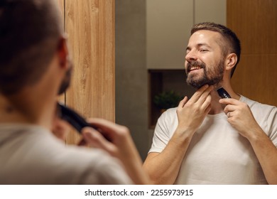 Reflection of a bearded man shaving his beard with shaver while standing at home in bathroom. - Shutterstock ID 2255973915