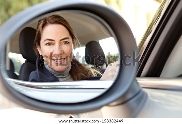 Reflection in a car reversing side\
mirror of a smart and mature woman driver, smiling at the camera\
while driving and arriving at home, feeling proud and\
happy.