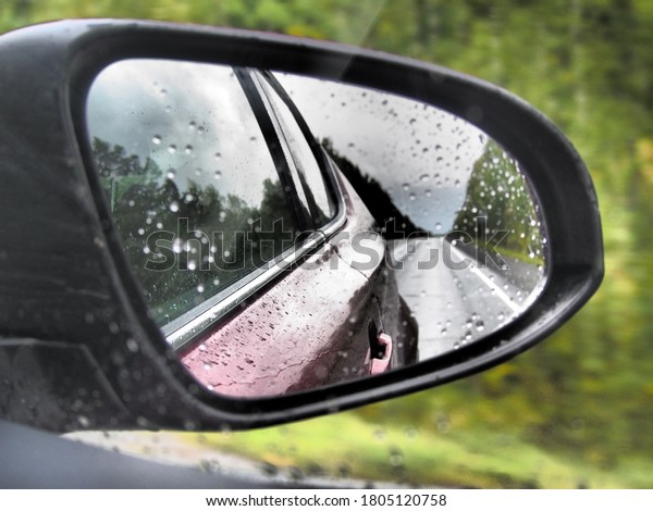 reflection of the forest and the\
road in the wet mirror of a red car while driving in rainy\
weather