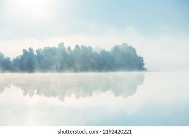 The reflection of forest trees in the still water of a foggy morning lake. Fog over dreamy autumn lake water.
