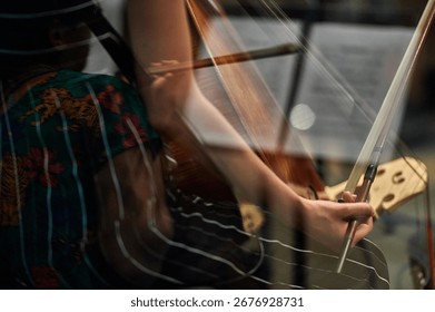 Reflection of a cellist’s hand holding a bow through glass, creating a layered composition that blends the musician’s movements with a colorful shirt in the background