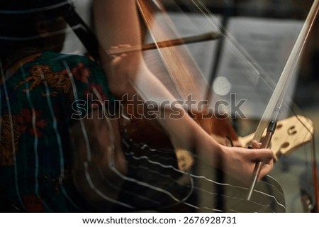 Reflection of a cellist’s hand holding a bow through glass, creating a layered composition that blends the musician’s movements with a colorful shirt in the background