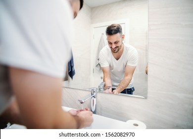 Reflection of handsome caucasian man standing in bathroom with cupped hands and washing himself in the morning.
